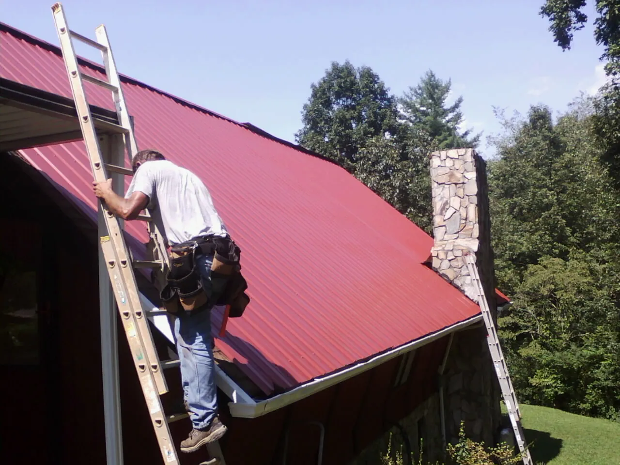 Roofer on a ladder beside a red metal roof and stone chimney — Soffit & Fascia Repair by Roofing Brothers in Wind Gap